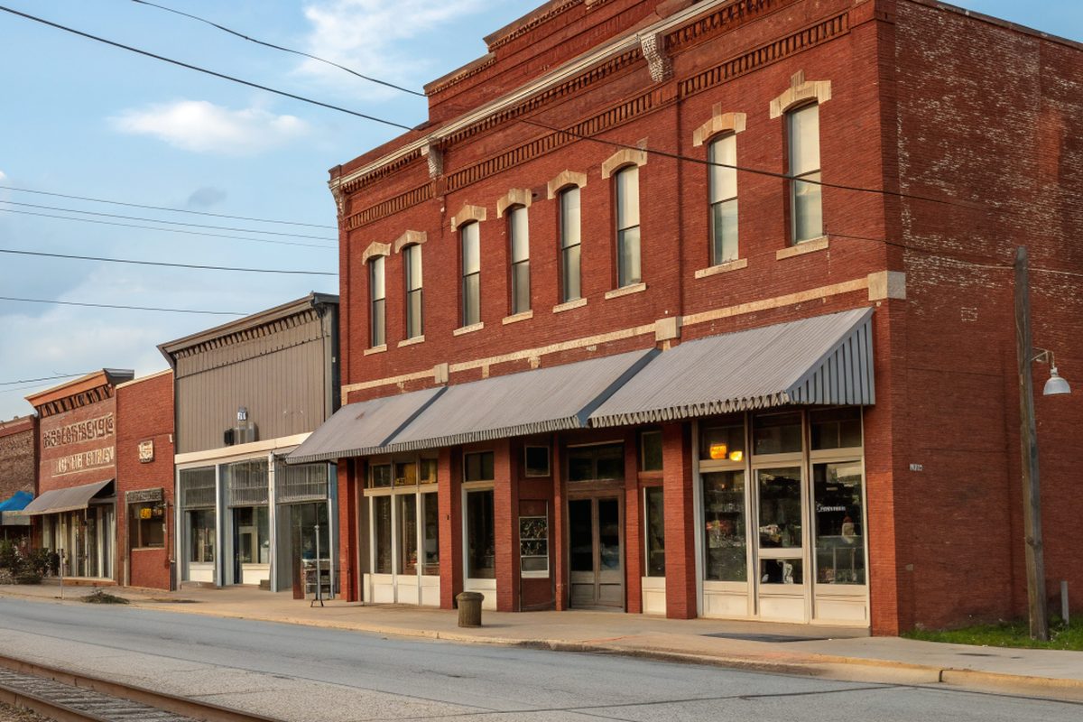 Old two-story red brick storefronts along a small-town main street beside railroad tracks